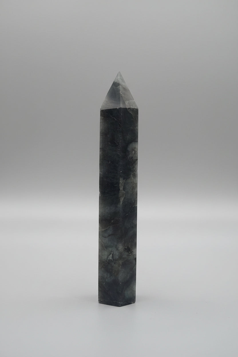 A dark gray natural quartz crystal obelisk with white and black veining, standing upright on a white surface against a textured gray wall.