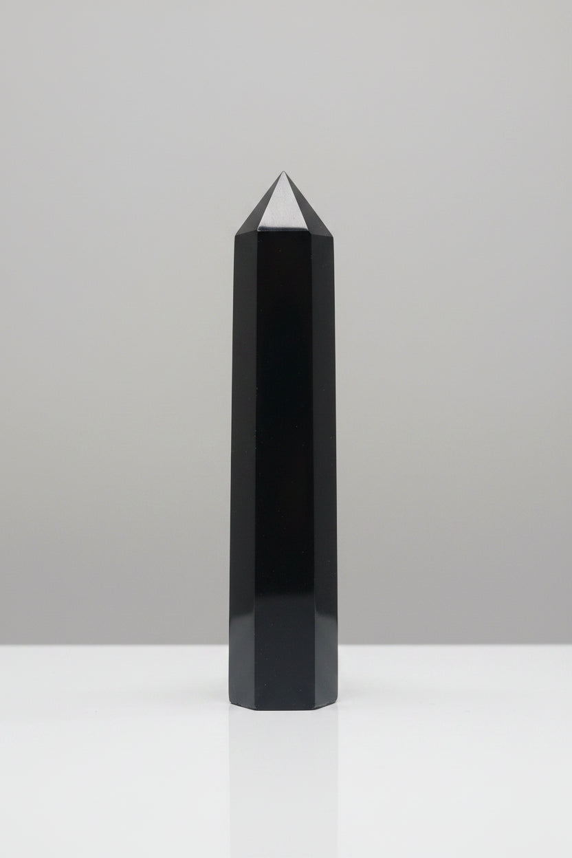 A tall, polished black crystal tower or obelisk standing upright on a white surface against a textured gray wall.